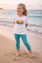 Girl wearing white sea turtle sunset kids t-shirt with turquoise leggings standing barefoot on sandy beach