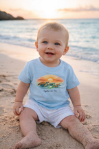 Baby wearing light blue turtle sunset t-shirt sitting on sandy beach – cute summer baby outfit