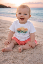 6 month old baby wearing white turtle sunset t-shirt and coral shorts sitting on sandy beach at sunset – cute summer baby outfit