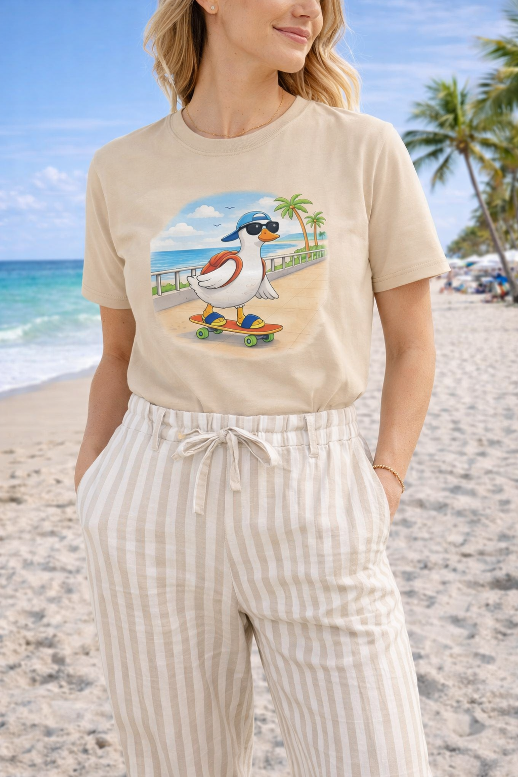 Woman wearing sand beige graphic t-shirt with cartoon duck skateboarding on a beach boardwalk, styled with striped pants in a sunny coastal setting