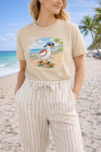 Woman wearing sand beige graphic t-shirt with cartoon duck skateboarding on a beach boardwalk, styled with striped pants in a sunny coastal setting